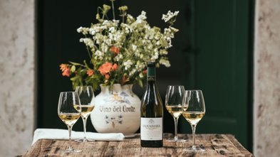 Ein rustikaler Holztisch trägt eine Flasche Vignasamara Wein und mehrere Gläser mit goldgelbem Wein, umgeben von frischen Blumen.