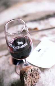 Ein Weinglas mit rotem Wein und dem Schriftzug 'OSBORNE' steht auf einem Holzuntergrund, umgeben von einem Korken und einer Etikette.