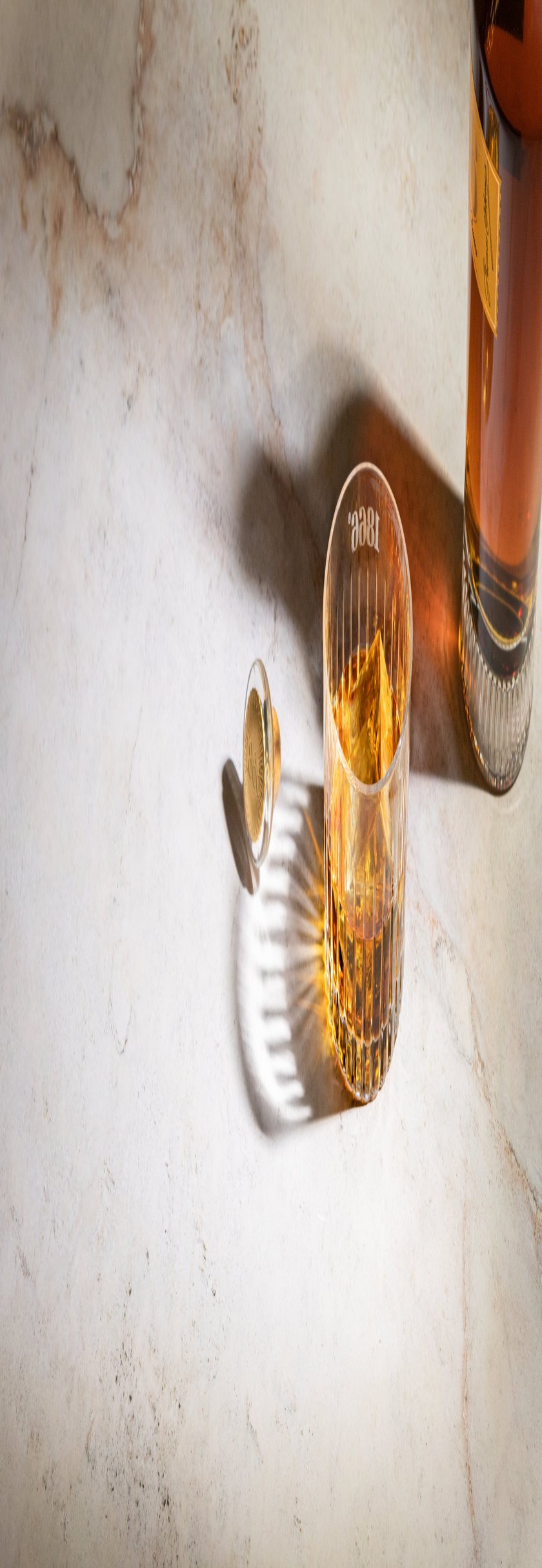 Ein Glas mit Whisky und Eiswürfeln wirft Schatten auf einen marmorierten Tisch, neben dem eine Flasche mit goldenem Getränk steht.