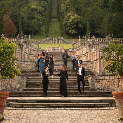 Eine Gruppe von elegant gekleideten Menschen geht eine steinerne Treppe hinunter, umgeben von üppigem Grün und historischen Statuen.