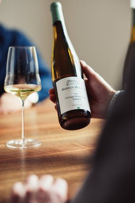 Eine Hand hält eine Flasche Riesling mit dem Etikett 'Markus Molitor', während ein Glas Wein auf dem Tisch steht.