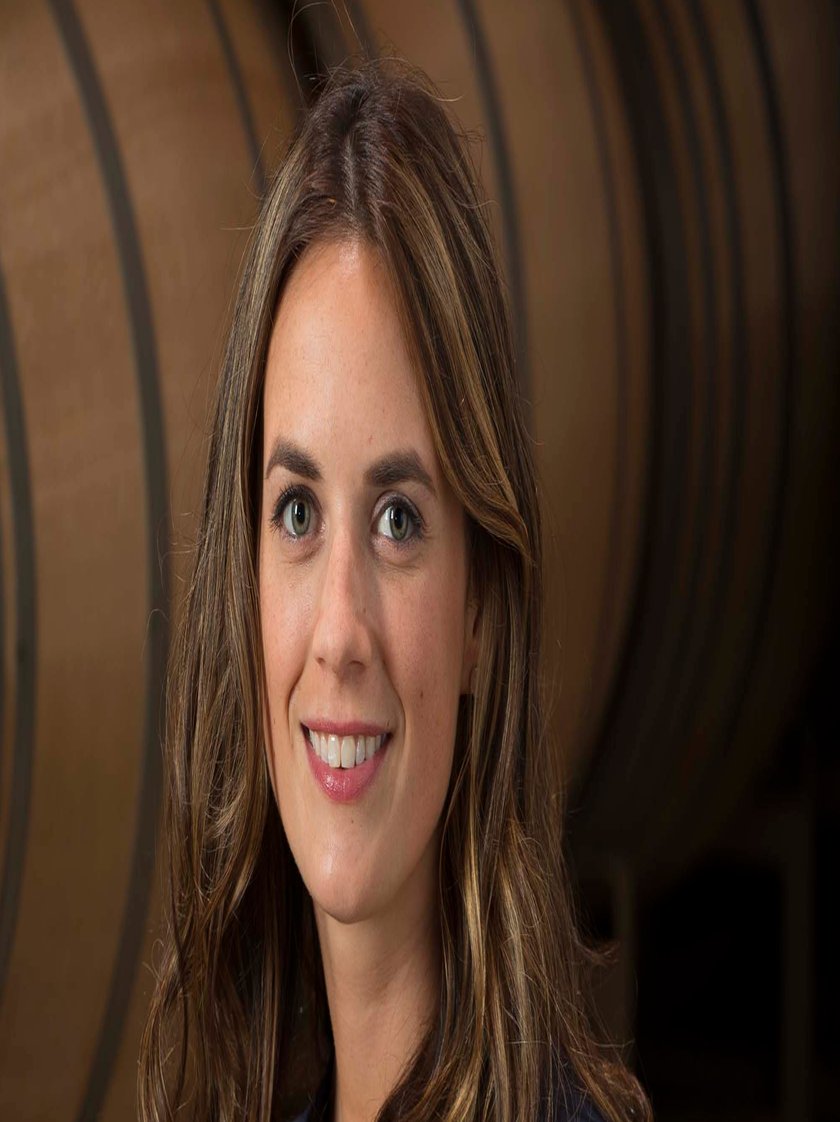 Eine Frau mit langen, lockigen Haaren lächelt, während sie ein Glas Rotwein in einem Weinkeller hält, umgeben von Holzfässern.