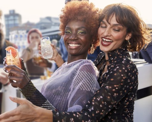 Zwei Frauen lachen fröhlich, während sie Cocktails in der Hand halten, umgeben von einer festlichen Atmosphäre.