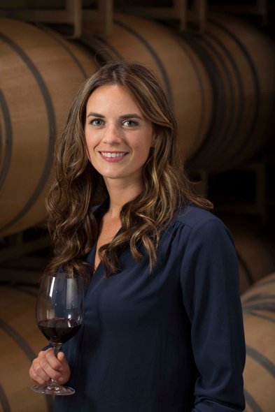 Eine Frau mit langen, lockigen Haaren lächelt, während sie ein Glas Rotwein in einem Weinkeller hält, umgeben von Holzfässern.