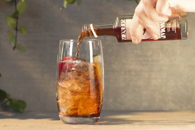 Ein Glas mit Eiswürfeln wird mit einem dunklen Getränk gefüllt, während frische Kräuter im Hintergrund sichtbar sind.