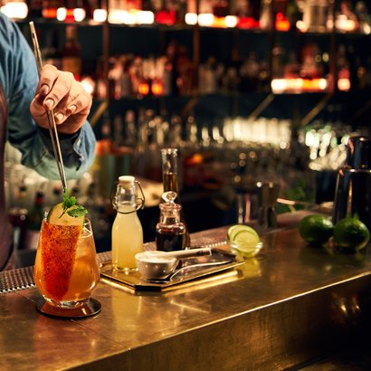 Ein Barkeeper in einem stilvollen Barambiente bereitet sorgfältig einen bunten Cocktail mit frischen Zutaten zu.