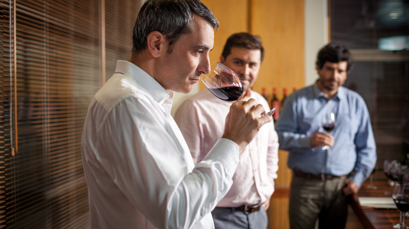 Ein Mann in einem weißen Hemd genießt einen Schluck Rotwein, während zwei andere Männer im Hintergrund aufmerksam zuschauen.