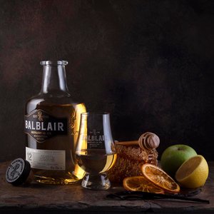 Eine Flasche Balblair Whisky steht neben einem Glas, umgeben von frischen Früchten und Honigwaben auf einem rustikalen Tisch.