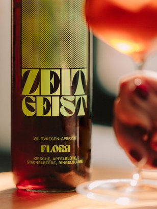 Eine Hand mit rotem Nagellack hält ein Glas, während eine Flasche 'ZEITGEIST' im Hintergrund sichtbar ist, die einen Aperitif präsentiert.