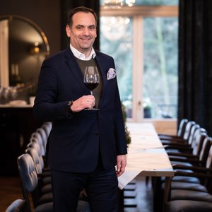 Der Mann in formeller Kleidung präsentiert stolz ein Weinglas, umgeben von einem langen Tisch und einer eleganten Atmosphäre.