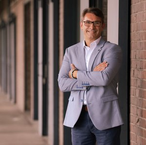 In einem eleganten Blazer und mit einem freundlichen Lächeln steht der Mann an einer Wand mit Fenstern.