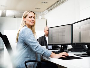 Eine blonde Frau sitzt an einem Schreibtisch mit zwei Monitoren, die Daten anzeigen, und lächelt freundlich in die Kamera.