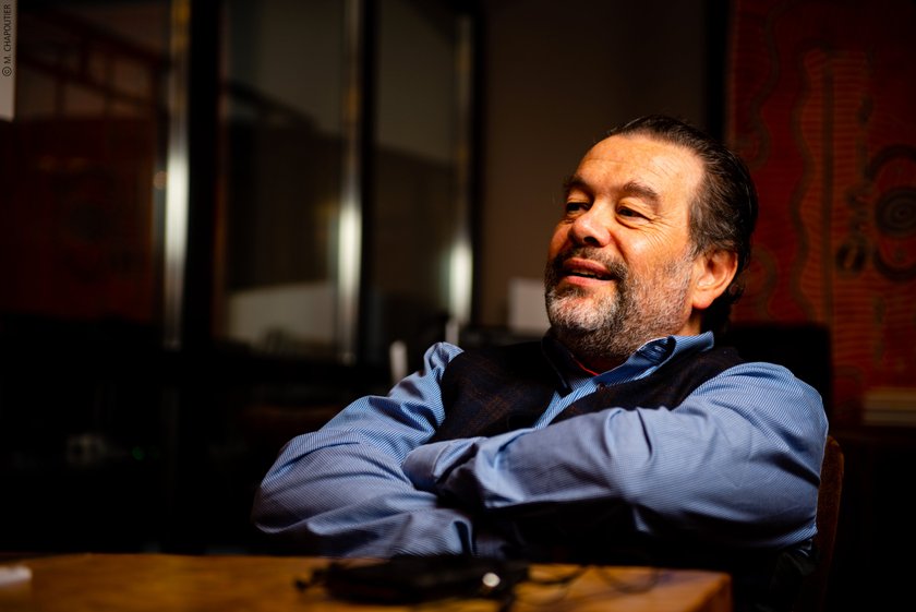 Ein lächelnder Mann mit Bart sitzt entspannt an einem Tisch, seine Arme sind verschränkt und er wirkt zufrieden.