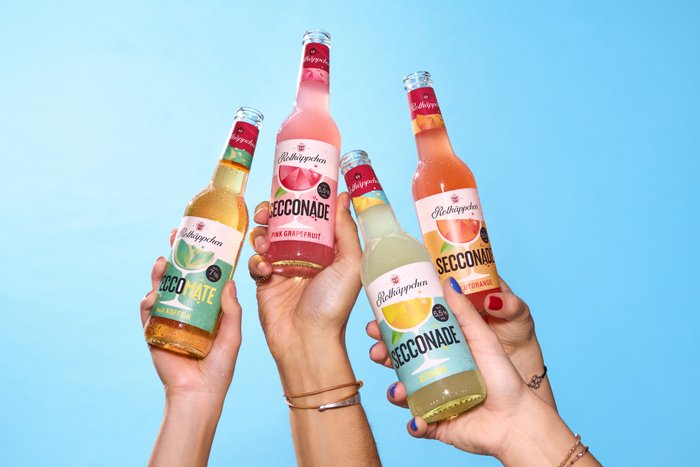 Vier Hände halten verschiedene Flaschen der Rotkäppchen-Secconade vor einem hellblauen Hintergrund, der Frische und Sommerlaune vermittelt.