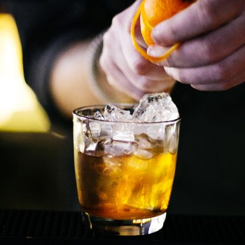 Ein Barkeeper drückt eine Orange über einem Glas mit Eis und einem goldenen Getränk aus, während die Bar im Hintergrund verschwommen ist.