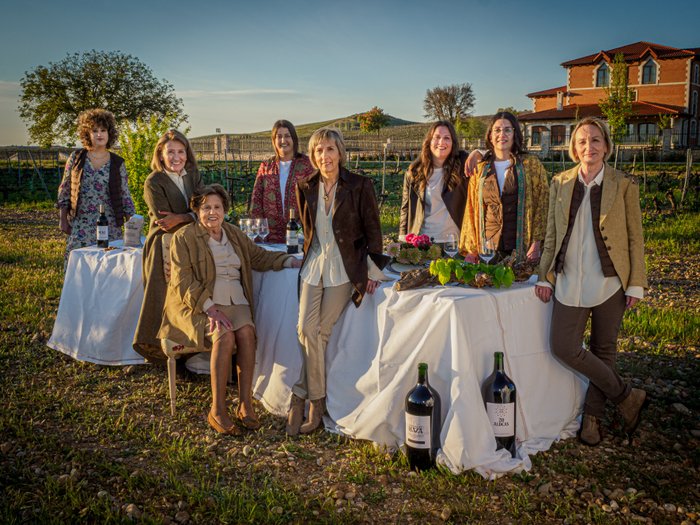 Eine Gruppe von acht Frauen steht in einem Weinberg, umgeben von Flaschen Wein und einem Tisch mit Gläsern und Blumen.