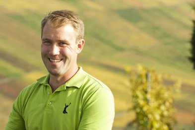 Ein lächelnder Mann in einem leuchtend grünen Poloshirt steht vor einer malerischen, hügeligen Landschaft mit sanften Farben.
