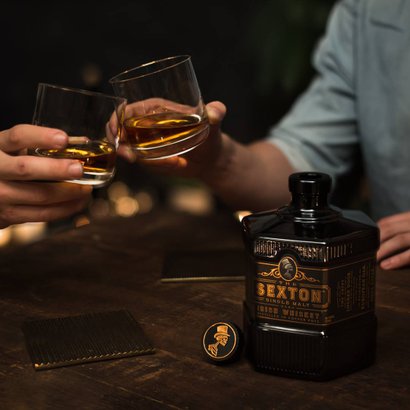 Zwei Hände stoßen mit Gläsern voll goldfarbenem Whiskey an, während eine Flasche Sexton Whiskey auf dem Tisch steht.