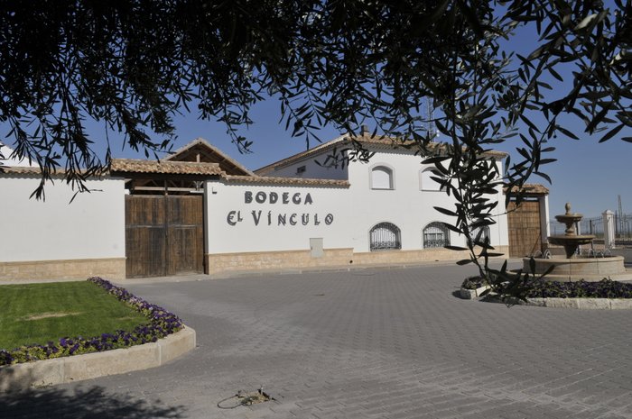 Die Fassade der Bodega el Vínculo zeigt eine elegante weiße Wand mit einem rustikalen Holztor und einem schönen Brunnen im Vordergrund.