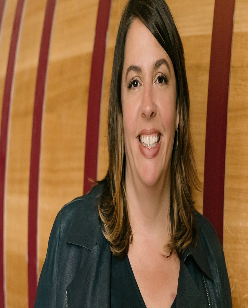 Vor einem großen Holzfass steht eine Frau mit langen, braunen Haaren, die in einer schwarzen Jacke lächelt.