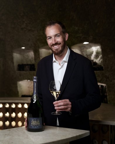 Ein lächelnder Mann in einem eleganten Anzug hält ein Glas Champagner und eine Flasche Charles Heidsieck in einer stilvollen Umgebung.