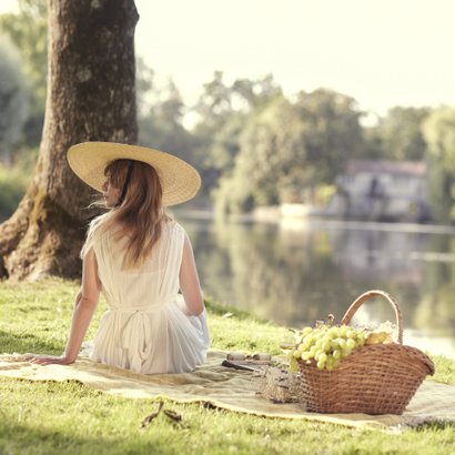 Die Szene zeigt eine friedliche Sommeratmosphäre, in der eine Frau in einem weißen Kleid am Wasser sitzt und die Aussicht bewundert.