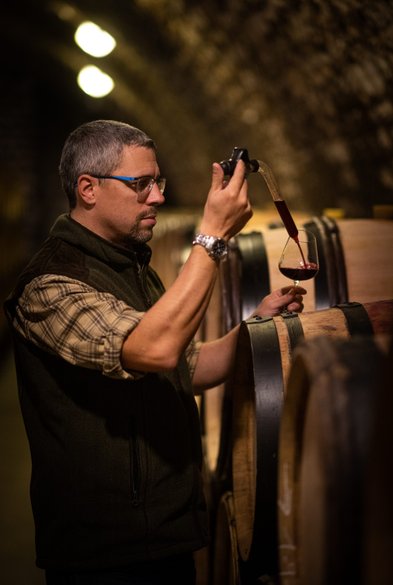 Ein Mann mit Brille und einem Hemd im Karomuster genießt den Moment, während er Wein aus einem Fass in ein Glas abfüllt.
