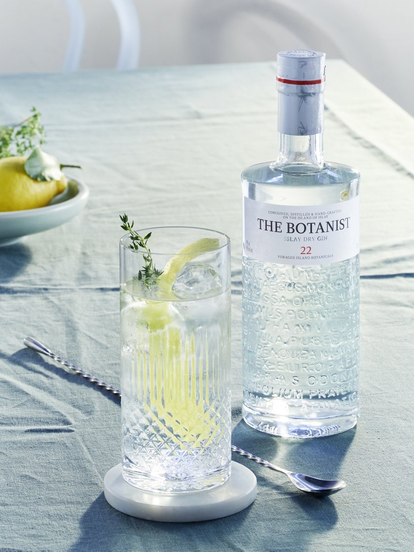 Ein erfrischender Gin-Cocktail mit Zitrone und Kräutern in einem Glas, das auf einem eleganten Tisch neben einer Flasche Botanist steht.