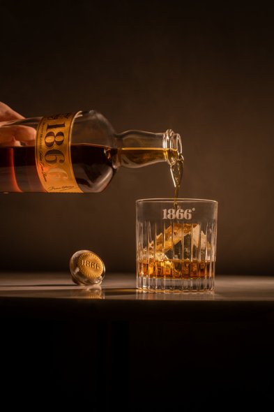 Ein elegantes Glas wird mit einem bernsteinfarbenen Getränk gefüllt, während die Flasche 1866 im sanften Licht glänzt.