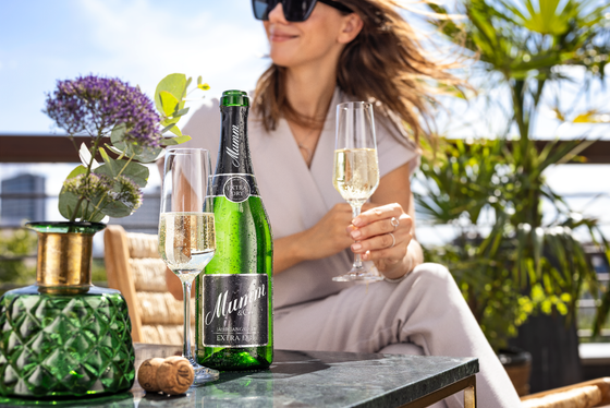 Eine Frau in Sonnenbrille genießt Sekt auf einer Terrasse, umgeben von Pflanzen und einem eleganten Tischarrangement.