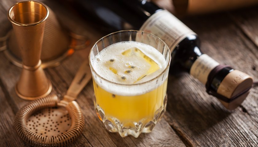 Ein Glas mit einem gelben Getränk, das mit Eiswürfeln gefüllt ist, steht auf einer rustikalen Holzoberfläche.