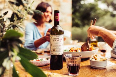 Auf dem Tisch sind bunte Salate und eine Flasche Rotwein zu sehen, während eine Person mit einem Löffel in einer Schüssel rührt.
