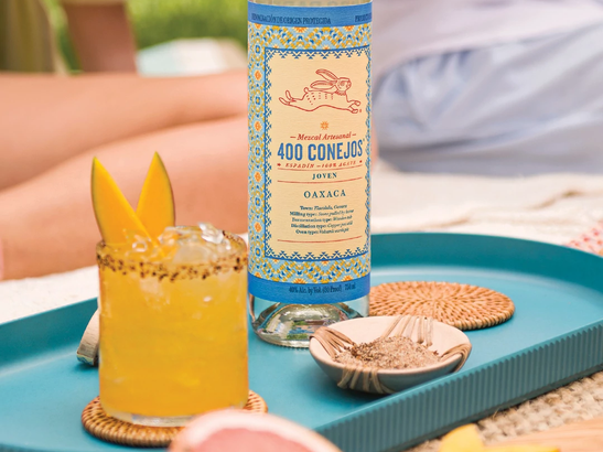 Ein erfrischendes Getränk mit Mangostücken und einer Zuckerrand-Dekoration steht auf einem blauen Tablett neben einer Flasche Mezcal.