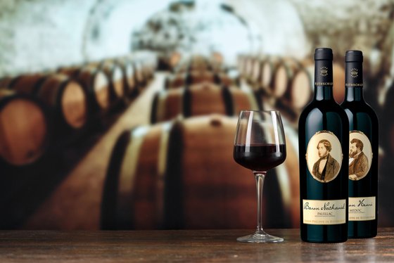 Ein elegantes Weinglas mit rotem Wein steht vor zwei Flaschen Wein, die in einem Weinkeller mit Holzfässern platziert sind.