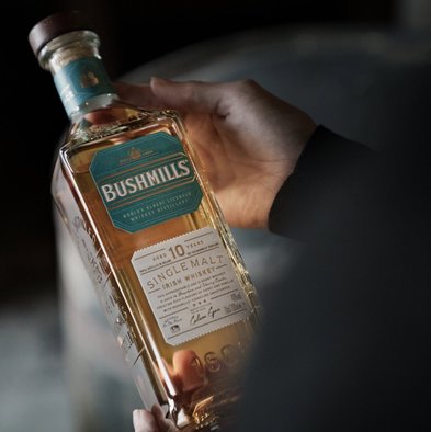 Eine Hand hält eine Flasche Bushmills 10 Jahre alten Single Malt Irish Whiskey, die in sanftem Licht glänzt.
