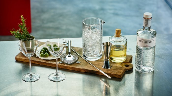 Zwei stilvolle Gläser und eine Auswahl an Oliven sind auf einem rustikalen Holzbrett für die Cocktailzubereitung angerichtet.