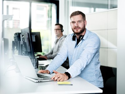 Ein Mann mit Bart und Kopfhörern steht an einem Schreibtisch und arbeitet an einem Laptop in einem modernen Büro.