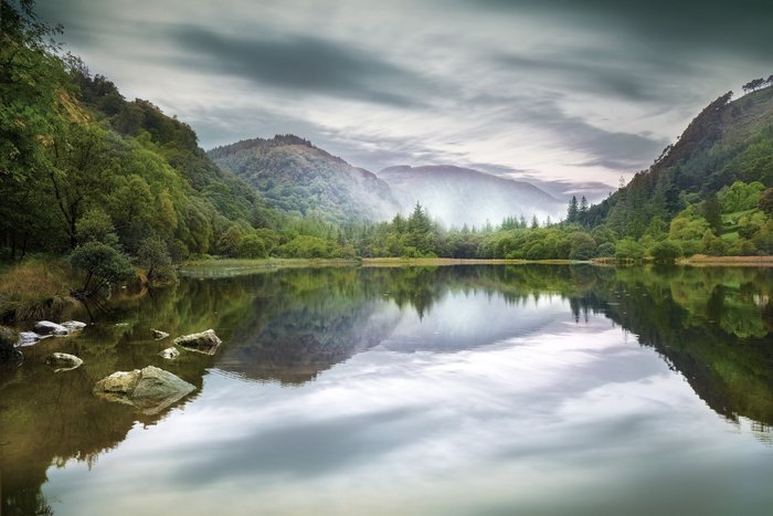 Ein ruhiger See spiegelt die sanften Hügel und bewaldeten Ufer wider, während der Himmel mit Wolken bedeckt ist.