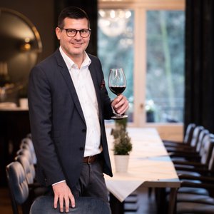 Der lächelnde Mann mit Brille präsentiert ein Glas Rotwein, während er in einem geschmackvoll eingerichteten Raum steht.