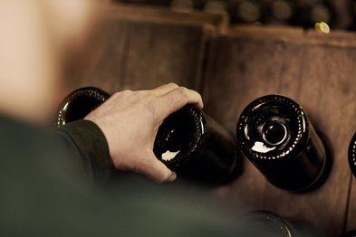 Eine Hand greift nach einer dunklen Flasche, während im Hintergrund weitere Flaschen auf einem Holzregal stehen.