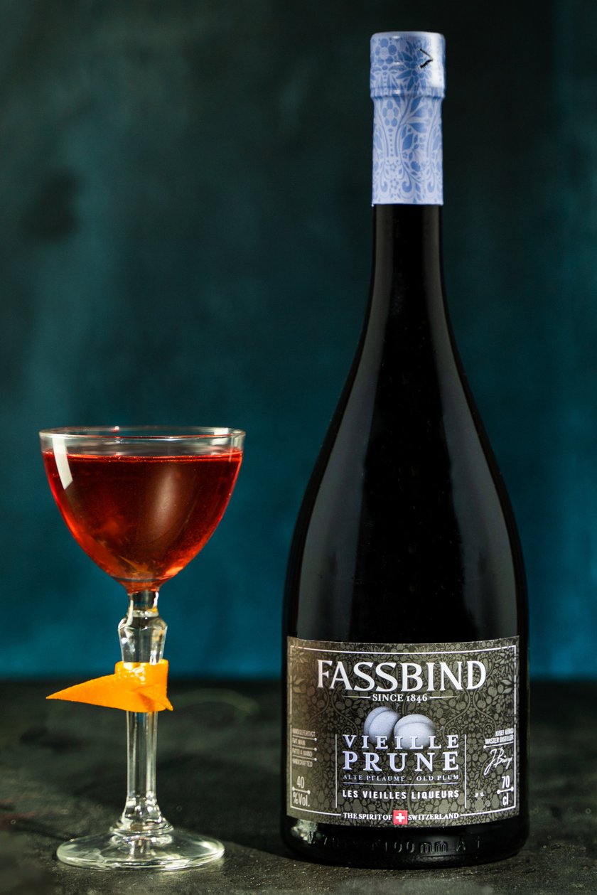 Ein elegantes Glas mit einem tiefroten Getränk, garniert mit einer Orangenschale, steht neben einer Flasche Fassbind Vieille Prune.