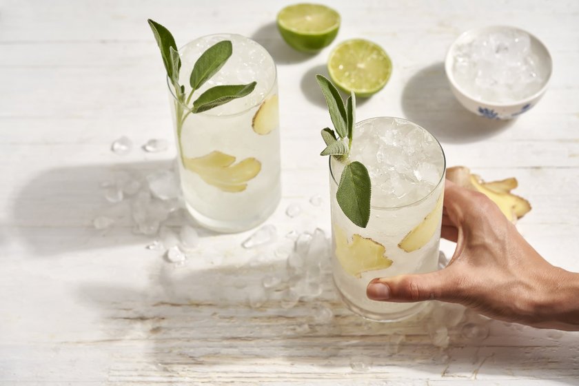 Zwei Gläser mit einem klaren Getränk, gefüllt mit Eis und Ingwer, stehen auf einem rustikalen Tisch, während Limetten im Hintergrund sichtbar sind.