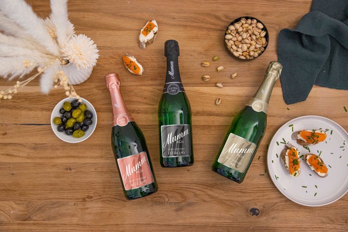 Drei Flaschen Mumm Sekt in verschiedenen Sorten liegen auf einem Holztisch, umgeben von Snacks und einer Vase mit Trockenblumen.