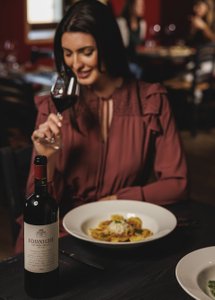 Eine Frau in einem eleganten, burgunderfarbenen Oberteil genießt ein Glas Rotwein und lächelt zufrieden.