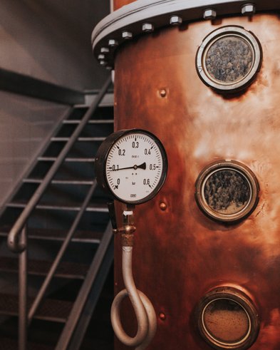 Ein Kupferkessel mit einem Manometer, das den Druck anzeigt, steht vor einer Treppe in einer Brauerei.