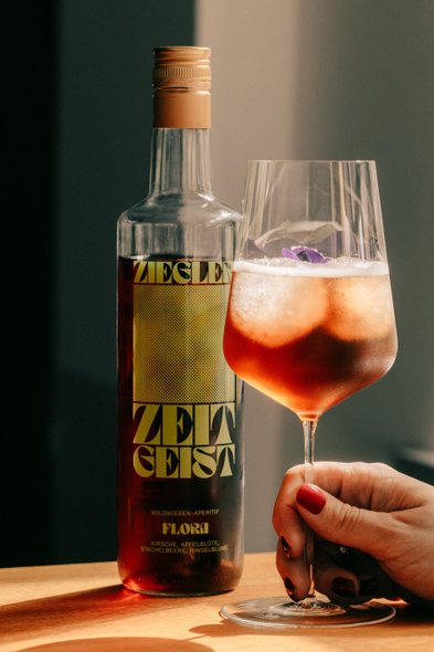 Ein erfrischendes Getränk in einem Glas mit einer Blüte, während die Flasche Zeitgeist-Aperitif auf einem Holzuntergrund steht.