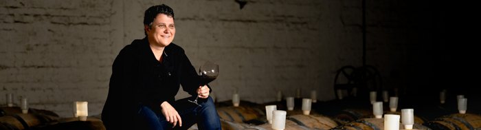 Ein fröhlicher Mann sitzt auf einem Weinfass und hält ein Glas Rotwein, umgeben von der rustikalen Kulisse eines Weinkellers.