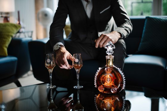 Ein elegant gekleideter Mann sitzt auf einem Sofa und hält ein Glas, während er eine edle Spirituosenflasche betrachtet.
