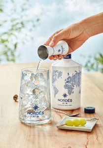 Eine Hand gießt Gin aus einem Messbecher in ein Glas mit Eiswürfeln, während eine Flasche Nordés Gin im Hintergrund steht.