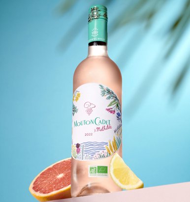 Vor einem sanften blauen Hintergrund präsentiert sich eine Flasche Mouton Cadet, dekoriert mit floralen Motiven.